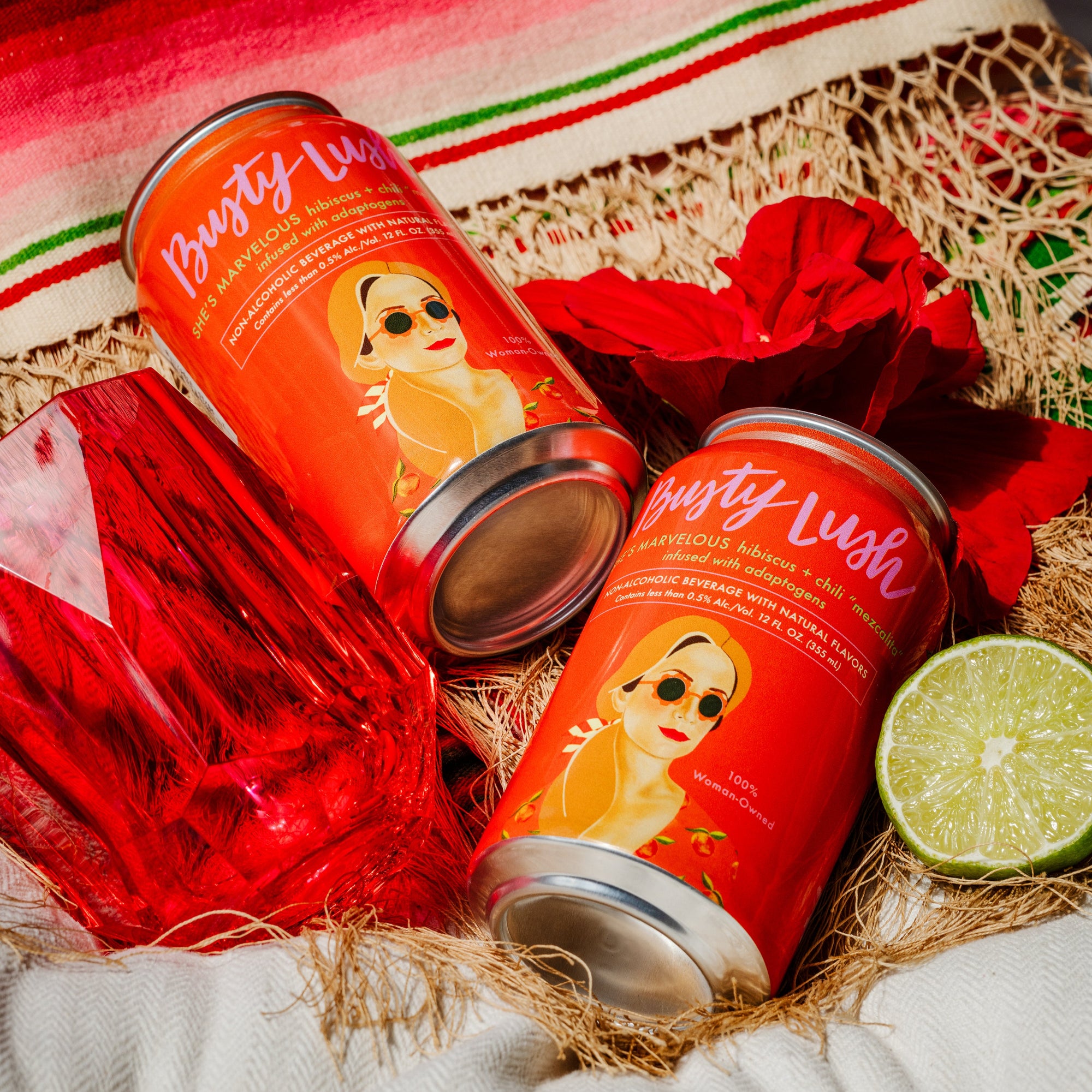 Two cans of 'Busty Lush' on a decorative surface with a colorful blanket and lime.